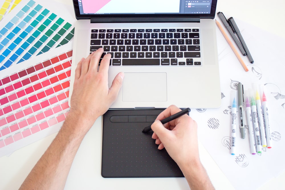 Hands drawing on a graphics tablet beside printed colour swatches and markers