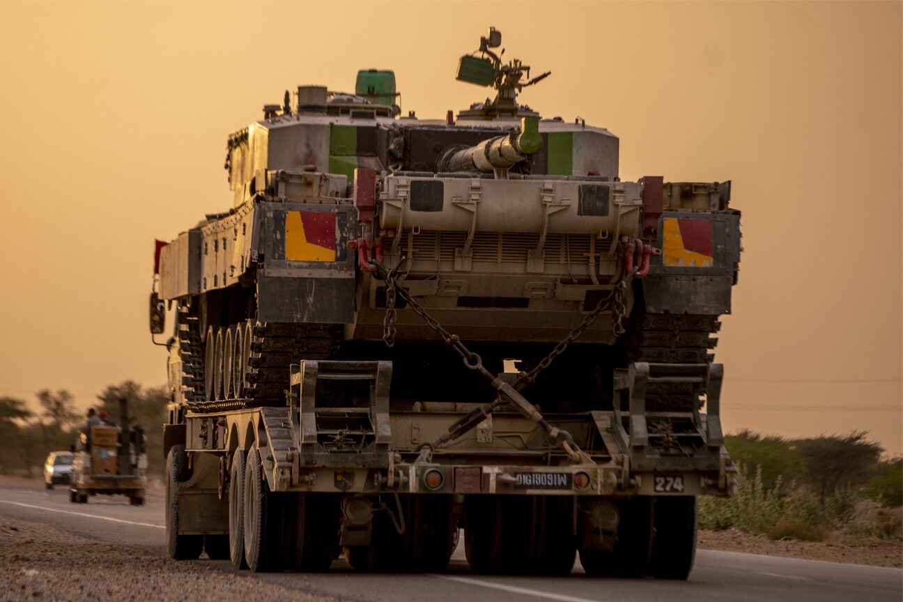 A tank chained down on a transporter trailer, seen from behind on a dusty road