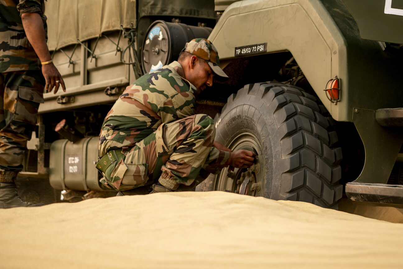 A soldier crouched in deep sand working on the rear wheel of a truck